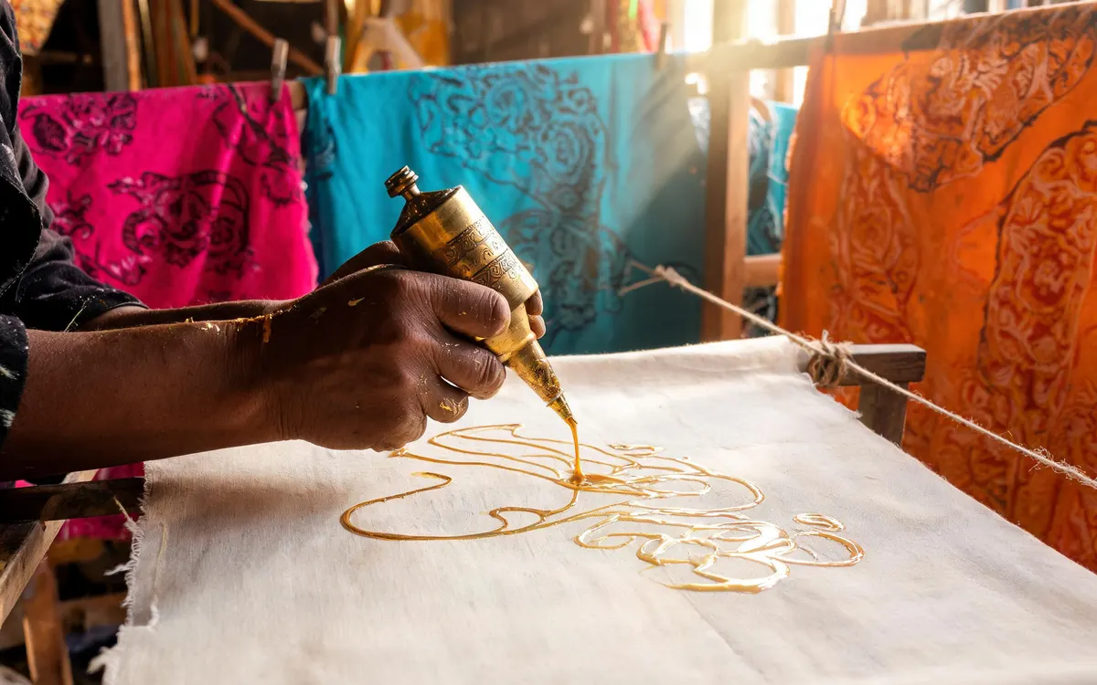 AI-generated website hero photo: artisan drawing wax patterns beside colourful batik fabric